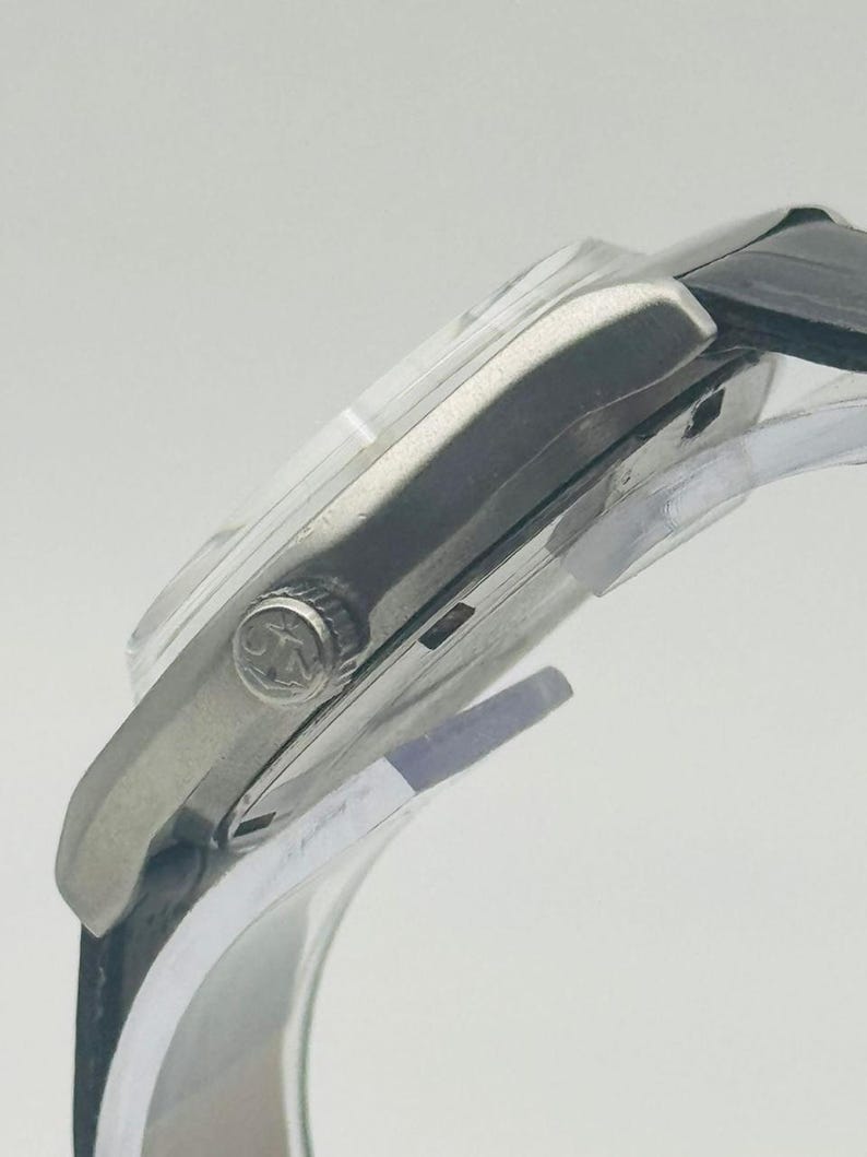 May include: A silver-toned wristwatch with a black leather band. The watch face is not visible, but the side profile shows a clear crystal and a textured crown. The watch is displayed on a clear stand.