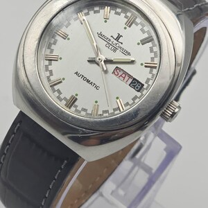 Puede incluir: Un reloj autom&aacute;tico Jaeger-LeCoultre Club plateado con esfera blanca. El reloj tiene una correa de cuero negro y muestra el d&iacute;a y la fecha. La esfera del reloj presenta el logotipo de Jaeger-LeCoultre y la palabra "AUTOMATIC".