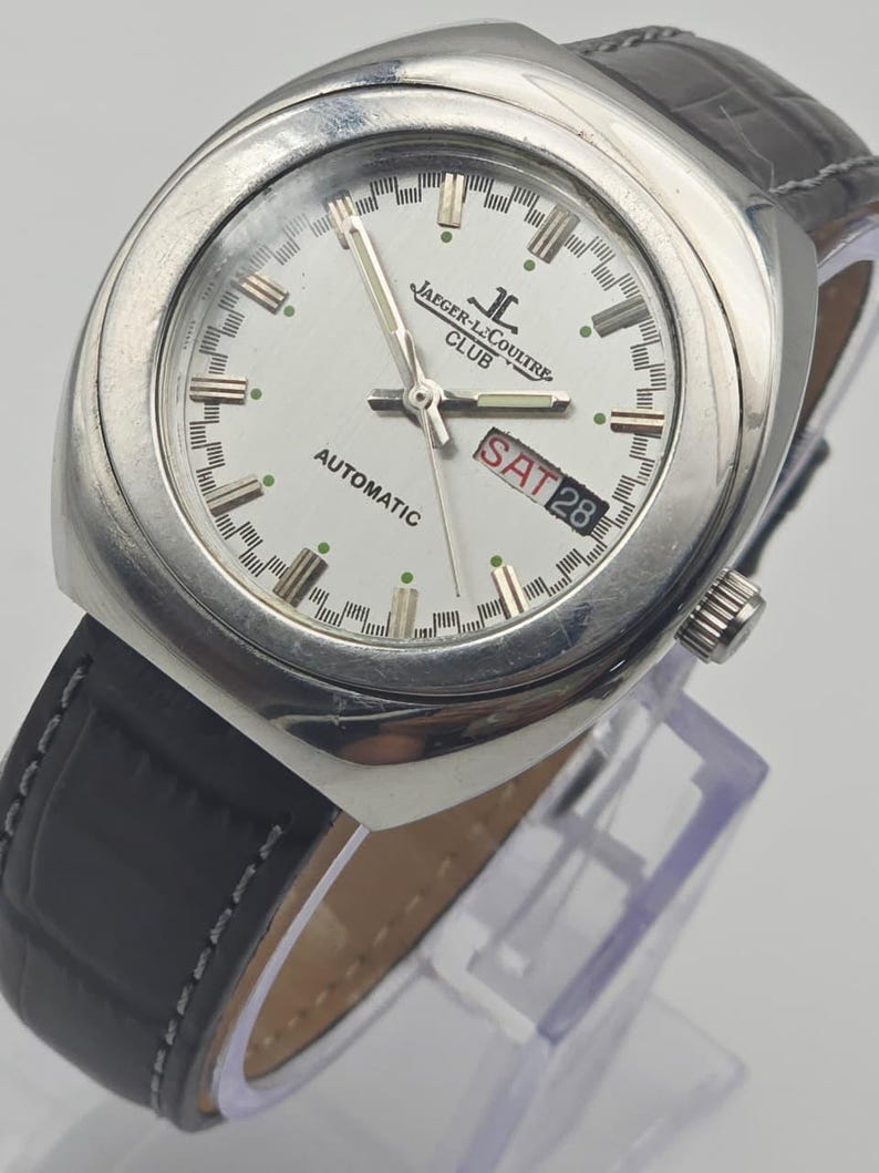 Puede incluir: Un reloj autom&aacute;tico Jaeger-LeCoultre Club plateado con esfera blanca. El reloj tiene una correa de cuero negro y muestra el d&iacute;a y la fecha. La esfera del reloj presenta el logotipo de Jaeger-LeCoultre y la palabra "AUTOMATIC".