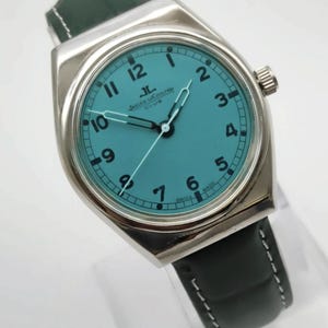 Pode incluir: Um rel&oacute;gio Jaeger-LeCoultre Club prateado com mostrador turquesa e ponteiros pretos. O rel&oacute;gio tem algarismos pretos e uma pulseira de couro preta com costuras brancas. O mostrador do rel&oacute;gio diz "JAEGER LECOULTRE CLUB".