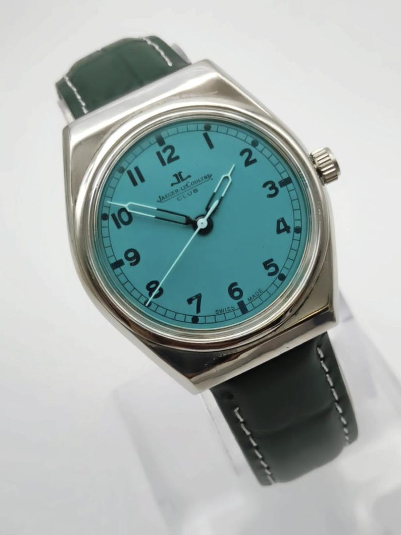 Pode incluir: Um rel&oacute;gio Jaeger-LeCoultre Club prateado com mostrador turquesa e ponteiros pretos. O rel&oacute;gio tem algarismos pretos e uma pulseira de couro preta com costuras brancas. O mostrador do rel&oacute;gio diz "JAEGER LECOULTRE CLUB".