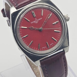 May include: A vintage Ulysse Nardin wristwatch with a red dial and silver-tone case. The watch features a burgundy leather strap and the brand's logo at the 12 o'clock position. The watch face has silver-tone hour markers and hands.