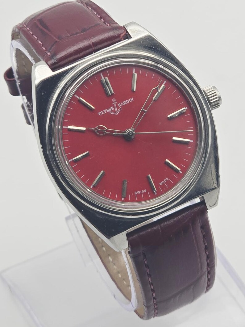 May include: A vintage Ulysse Nardin wristwatch with a red dial and silver-tone case. The watch features a burgundy leather strap and the brand's logo at the 12 o'clock position. The watch face has silver-tone hour markers and hands.
