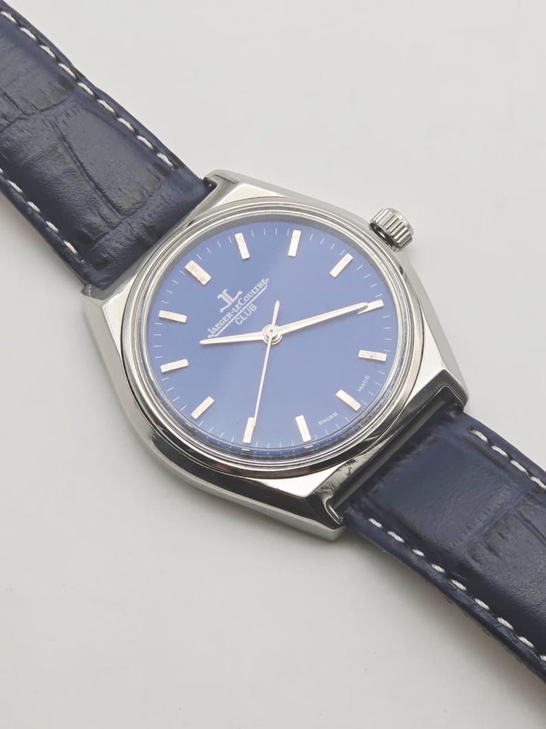 May include: A silver-toned wristwatch with a blue face and a navy blue leather band. The watch face features silver hour markers and hands. The band is stitched with white thread. The watch is a classic timepiece.