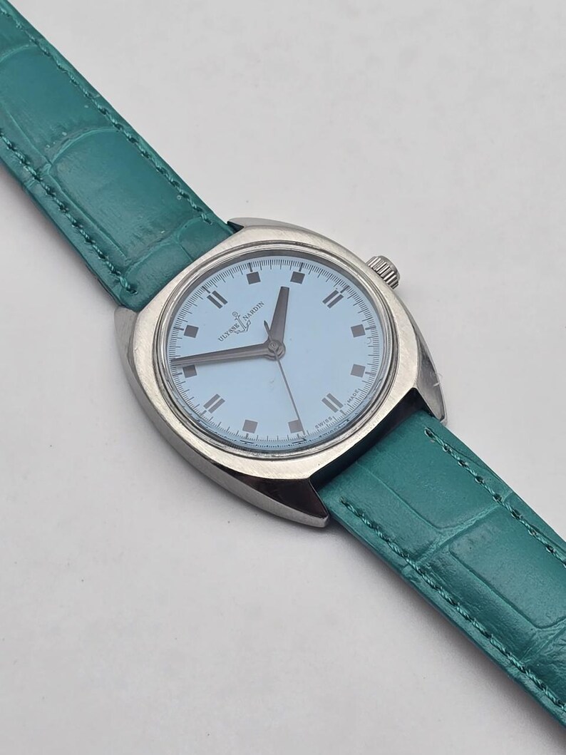Peut inclure: Une montre-bracelet argent&eacute;e avec un cadran bleu clair et un bracelet en cuir vert d'eau. Le cadran affiche la marque "Ulysse Nardin" et des rep&egrave;res horaires simples. Le bracelet pr&eacute;sente un motif textur&eacute;, semblable &agrave; du crocodile.