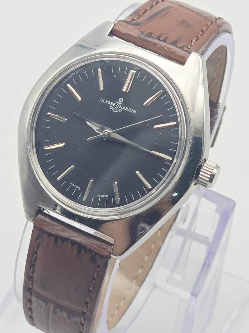 May include: A silver-toned Ulysse Nardin wristwatch with a black face and brown leather band. The watch face has gold-colored markers and hands. The band is a dark brown, and the watch is displayed on a clear stand.