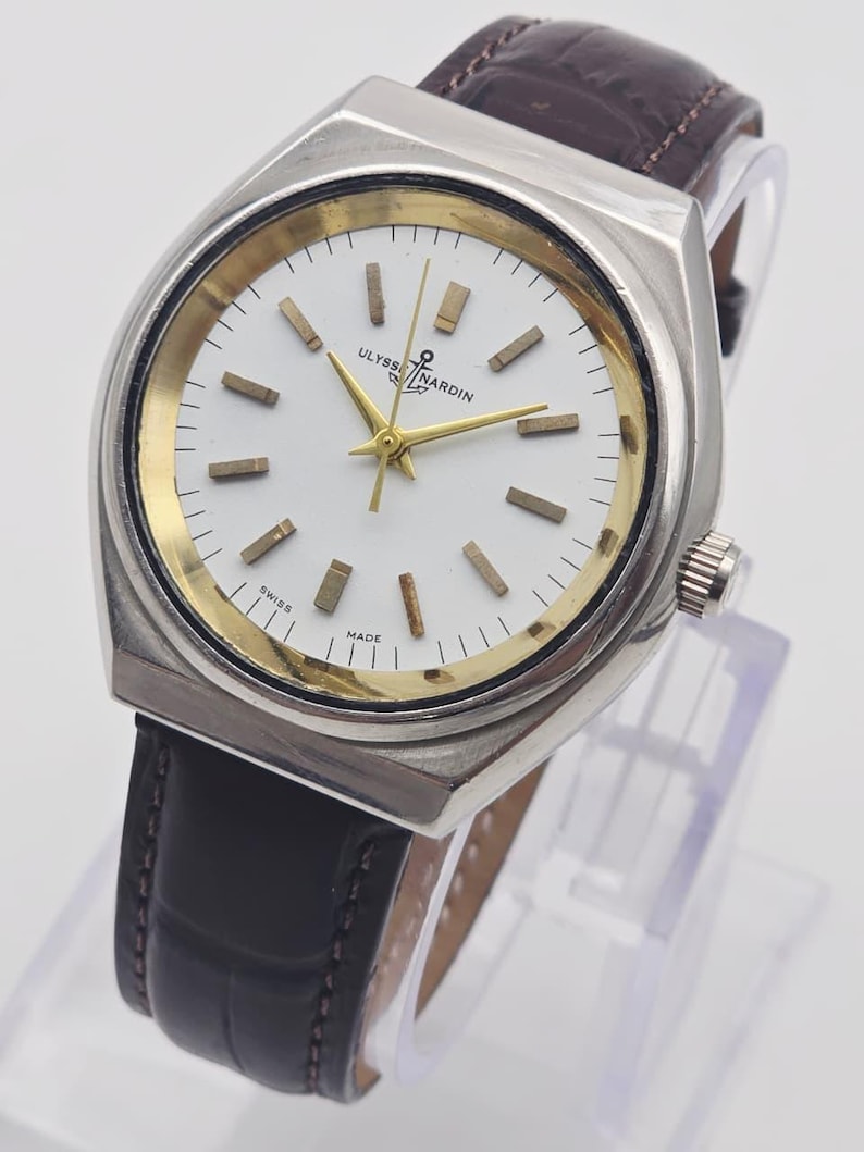 Pu&ograve; includere: Un orologio Ulysse Nardin vintage con cassa color argento, quadrante bianco con dettagli dorati e cinturino in pelle marrone. Il quadrante dell'orologio presenta lancette e indici dorati. L'orologio &egrave; marcato "SWISS MADE".