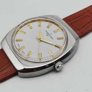 May include: A silver-toned Ulysse Nardin wristwatch with a white face and gold-colored hands and markers. The watch has a brown leather strap and a square-shaped case. The watch face displays the brand name and "Swiss Made".