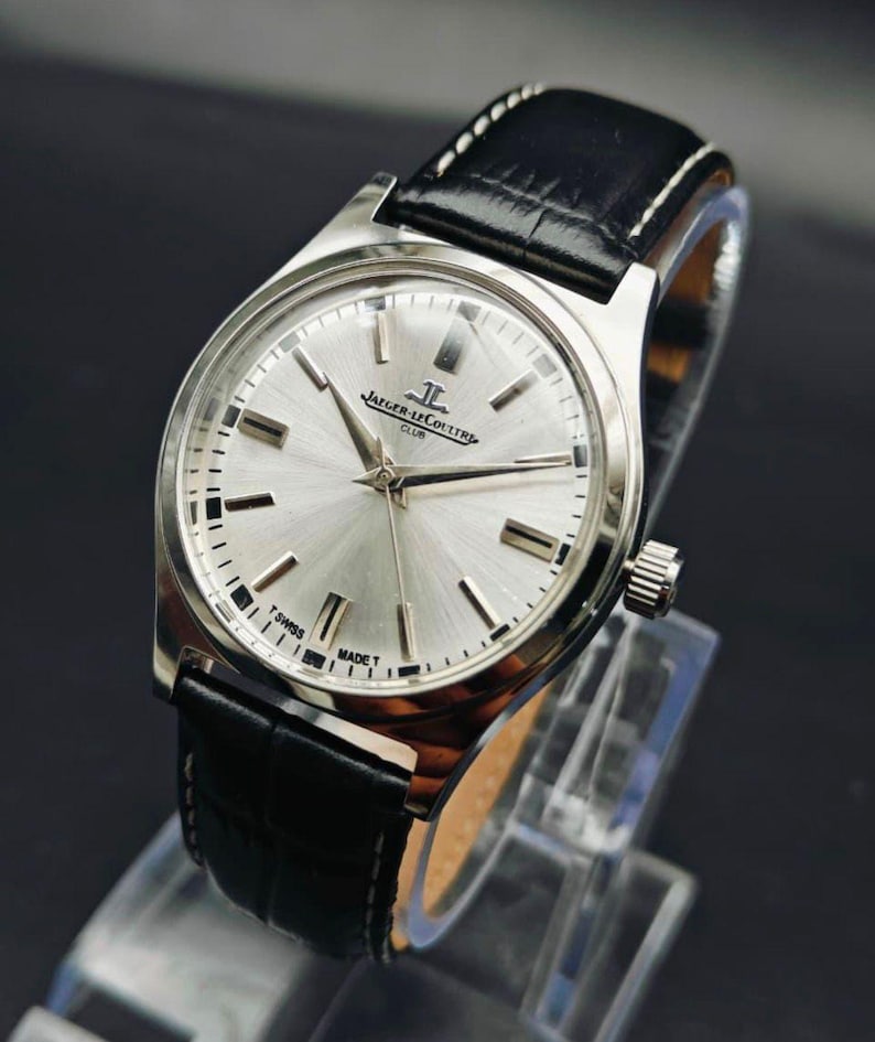 May include: A silver-toned Jaeger-LeCoultre Club watch with a black leather band. The watch face is silver with black hands and markers. The watch has a crown on the right side and is displayed on a clear stand.