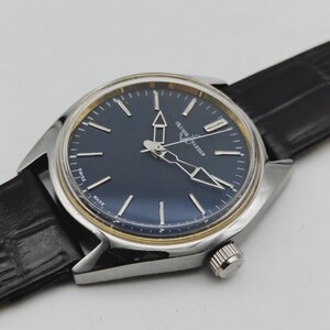 May include: A silver-toned wristwatch with a dark blue face and gold-colored accents. The watch features silver hands and markers, a black leather band, and a textured crown. The watch face has the text "Ulysse Nardin" and "Swiss Made".