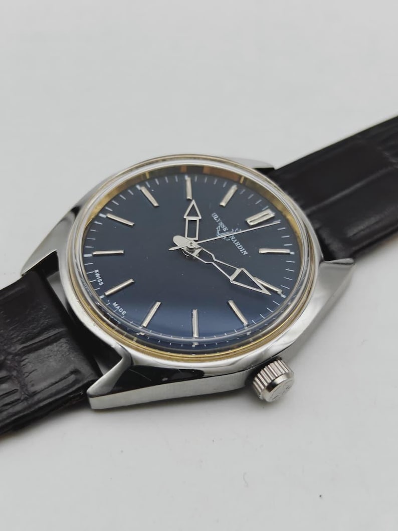 May include: A silver-toned wristwatch with a dark blue face and gold-colored accents. The watch features silver hands and markers, a black leather band, and a textured crown. The watch face has the text "Ulysse Nardin" and "Swiss Made".