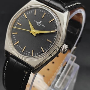 May include: A Ulysse Nardin wristwatch with a round black dial, gold-tone hands, and gold-tone hour markers. The watch features a silver-tone metal case and a black leather strap with white stitching. The text Ulysse Nardin and Swiss Made are visible.