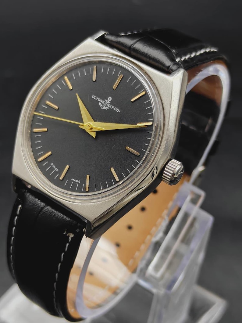 May include: A Ulysse Nardin wristwatch with a round black dial, gold-tone hands, and gold-tone hour markers. The watch features a silver-tone metal case and a black leather strap with white stitching. The text Ulysse Nardin and Swiss Made are visible.