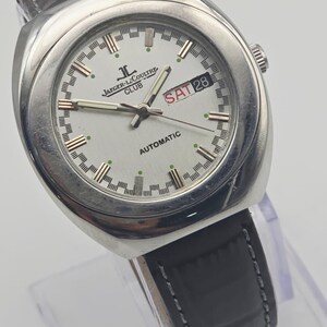 Puede incluir: Un reloj autom&aacute;tico Jaeger-LeCoultre Club plateado con esfera blanca. El reloj tiene una correa de cuero negro y muestra "SAT 28" en un recuadro rojo. La esfera redonda tiene detalles dorados y verdes.
