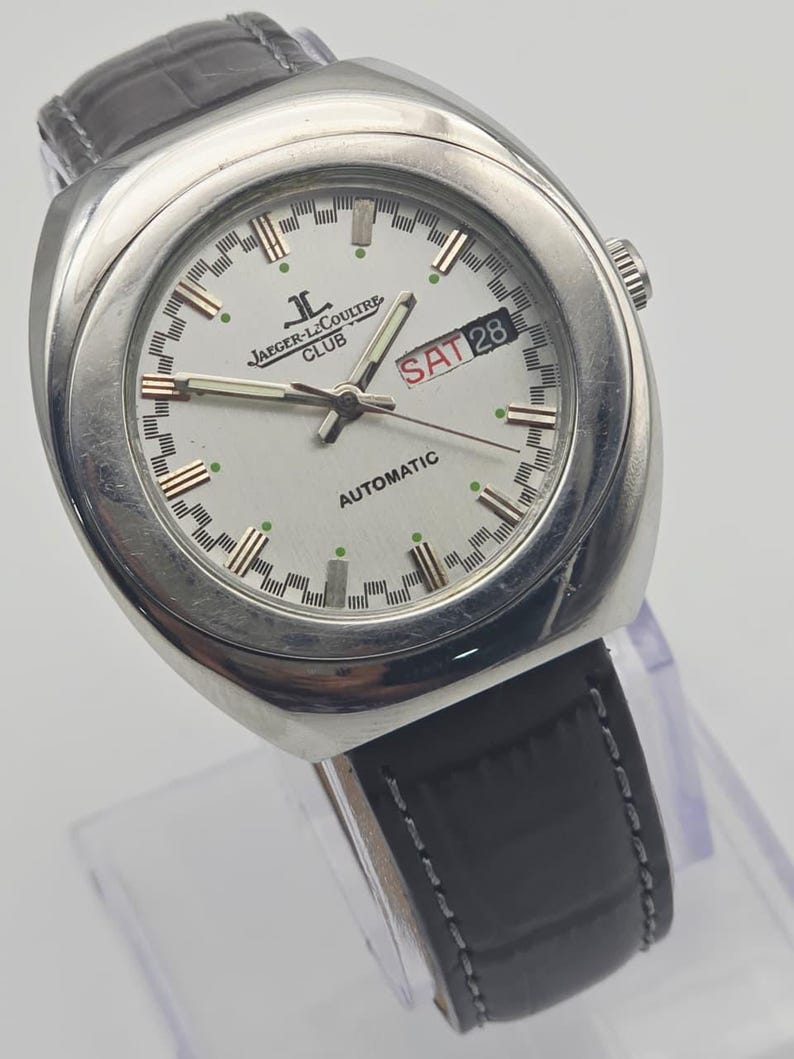Puede incluir: Un reloj autom&aacute;tico Jaeger-LeCoultre Club plateado con esfera blanca. El reloj tiene una correa de cuero negro y muestra "SAT 28" en un recuadro rojo. La esfera redonda tiene detalles dorados y verdes.