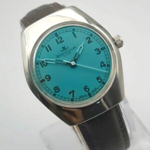 Op de afbeelding: Een vintage Jaeger-LeCoultre Club horloge met een turquoise wijzerplaat en zilveren kast. Het horloge heeft zwarte cijfers, zilveren wijzers en een bruine leren band. Het horloge is gemarkeerd met "Swiss Made".