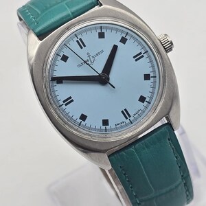 Peut inclure: Une montre-bracelet vintage Ulysse Nardin avec un cadran bleu clair et des aiguilles et marqueurs noirs. Le bo&icirc;tier est argent&eacute; et le bracelet en cuir est de couleur teal. Le cadran porte les inscriptions "Ulysse Nardin" et "Swiss Made".