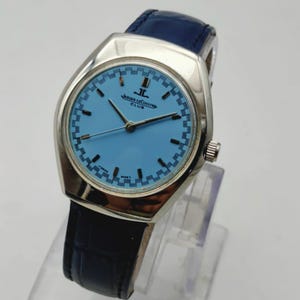 May include: A silver Jaeger-LeCoultre Club watch with a light blue face and a dark blue leather strap. The watch face has a checkered pattern around the edge and the text "JAEGER-LECOULTRE CLUB".