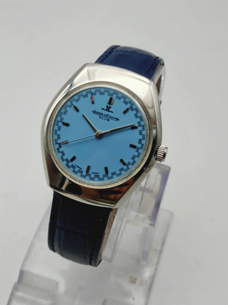 May include: A silver Jaeger-LeCoultre Club watch with a light blue face and a dark blue leather strap. The watch face has a checkered pattern around the edge and the text "JAEGER-LECOULTRE CLUB".