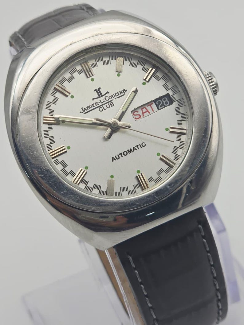 Puede incluir: Un reloj autom&aacute;tico Jaeger-LeCoultre Club plateado con esfera blanca, correa de cuero negro y visualizaci&oacute;n del d&iacute;a y la fecha. La esfera del reloj presenta el logotipo de Jaeger-LeCoultre y la palabra "AUTOMATIC". La fecha indica "SAB 28".