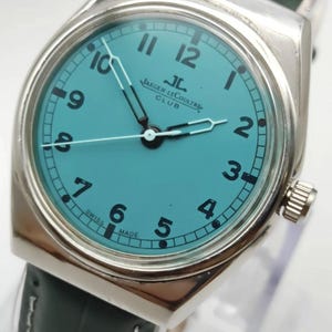 Pode incluir: Um rel&oacute;gio Jaeger-LeCoultre Club prateado com mostrador turquesa e algarismos pretos. O rel&oacute;gio tem uma bracelete de couro verde e uma moldura prateada. O mostrador exibe o texto "Jaeger-LeCoultre Club" e "Swiss Made".
