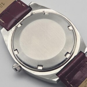 May include: A silver-toned wristwatch with a burgundy leather strap. The watch case is rectangular with a circular back. The strap is a deep burgundy color with a lighter tan underside. The watch face is not visible.