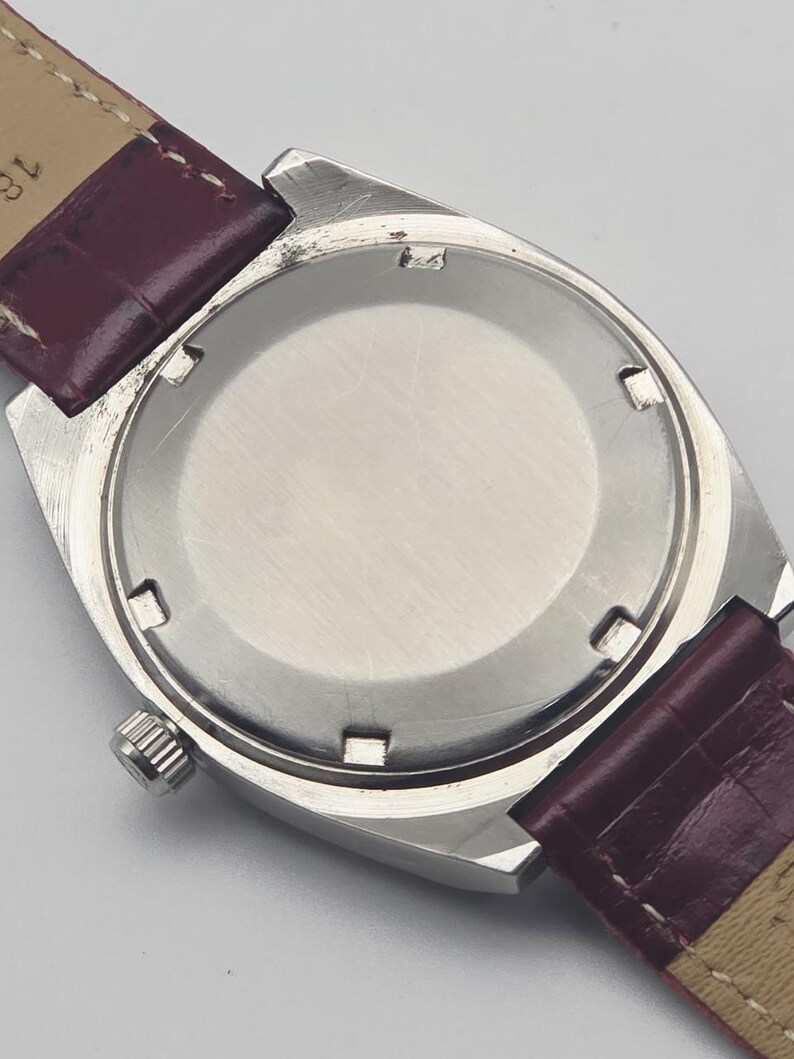 May include: A silver-toned wristwatch with a burgundy leather strap. The watch case is rectangular with a circular back. The strap is a deep burgundy color with a lighter tan underside. The watch face is not visible.