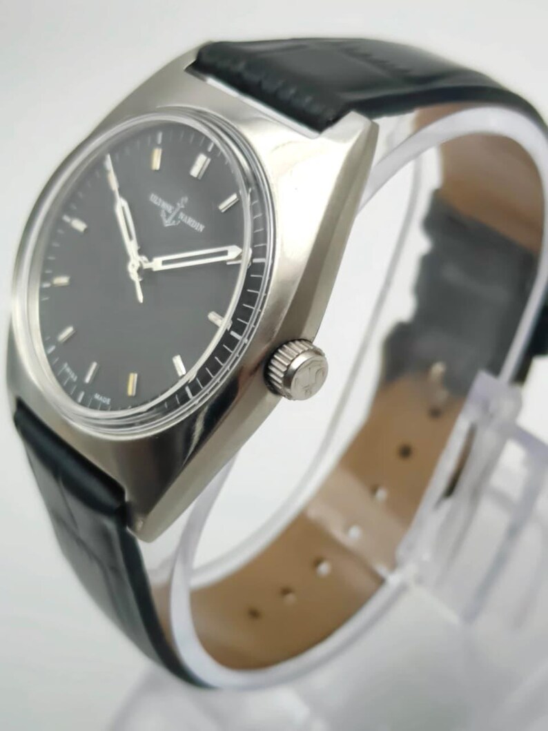 Puede incluir: Un reloj de pulsera plateado con esfera negra y correa de cuero negro. El reloj tiene una caja rectangular y una corona plateada. La esfera tiene agujas e indicadores plateados. La correa mide unos 20 cm.