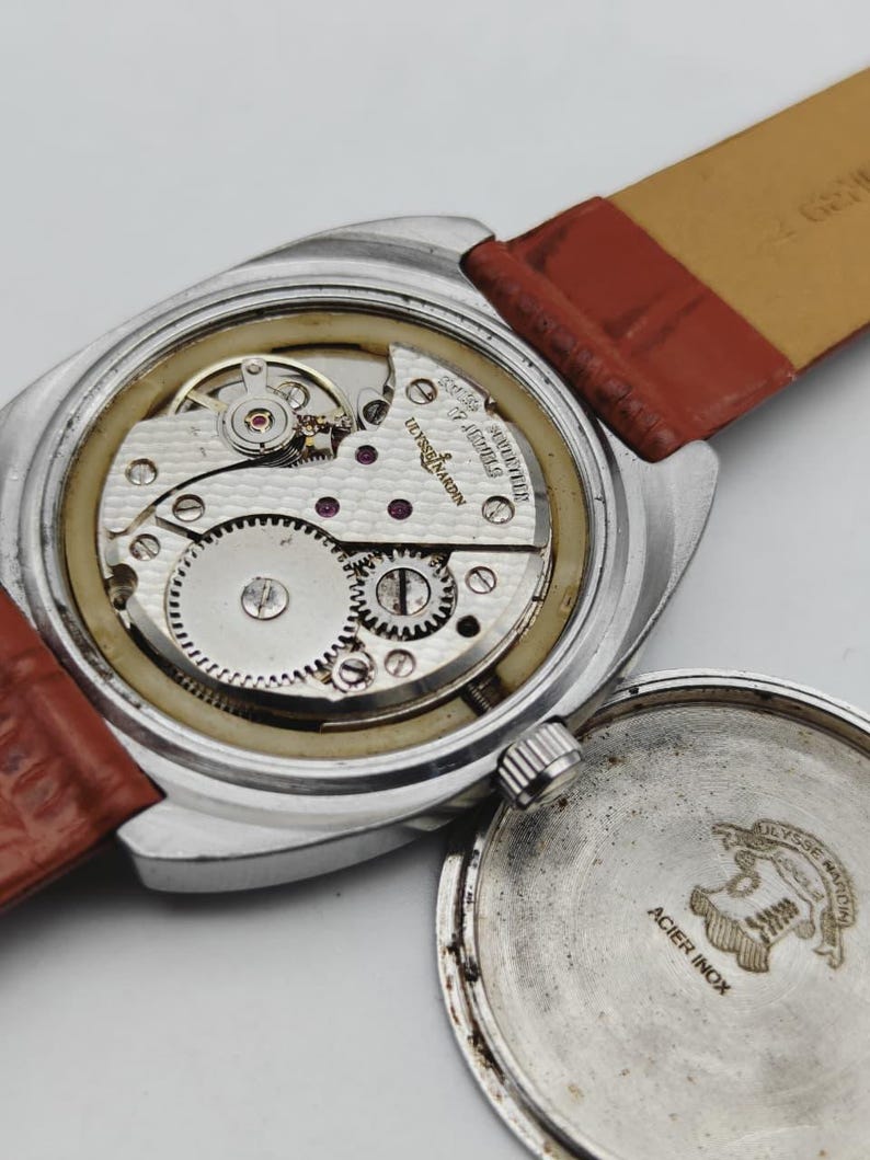 May include: Close-up of a vintage Ulysse Nardin watch with the back removed, revealing the intricate mechanical movement. The watch has a silver case and a brown leather strap. The words "ACIER INOX" are visible on the back.