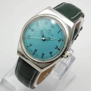 Pode incluir: Um rel&oacute;gio prateado com mostrador turquesa e pulseira de couro verde escuro. O rel&oacute;gio apresenta uma caixa retangular, algarismos brancos e o texto "Jaeger-LeCoultre Club". A pulseira &eacute; verde escuro com costuras brancas.
