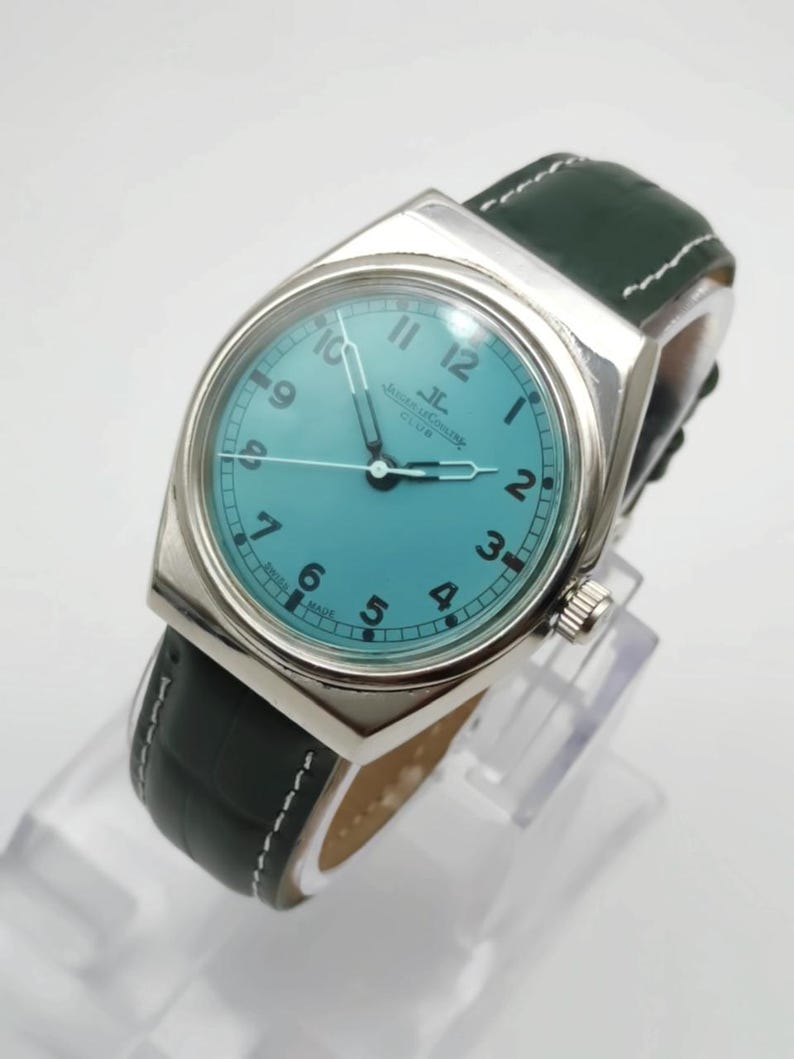 Pode incluir: Um rel&oacute;gio prateado com mostrador turquesa e pulseira de couro verde escuro. O rel&oacute;gio apresenta uma caixa retangular, algarismos brancos e o texto "Jaeger-LeCoultre Club". A pulseira &eacute; verde escuro com costuras brancas.