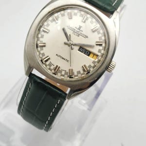 Puede incluir: Un reloj autom&aacute;tico Jaeger-LeCoultre Club plateado con esfera blanca y correa de cuero verde. La esfera muestra el d&iacute;a de la semana y el nombre de la marca. El reloj se exhibe sobre un soporte transparente.