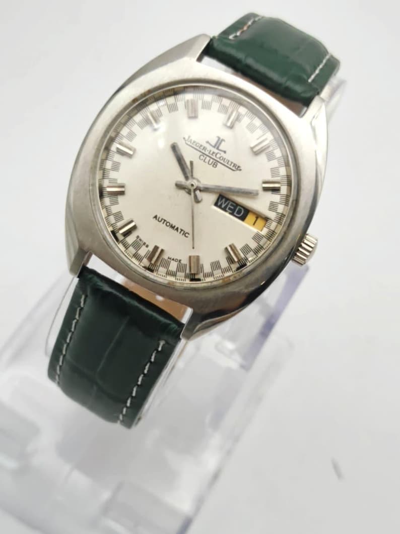Puede incluir: Un reloj autom&aacute;tico Jaeger-LeCoultre Club plateado con esfera blanca y correa de cuero verde. La esfera muestra el d&iacute;a de la semana y el nombre de la marca. El reloj se exhibe sobre un soporte transparente.
