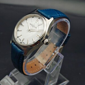 Peut inclure: Une montre-bracelet argent&eacute;e avec un cadran blanc et un bracelet en cuir bleu. La montre a un cadran rond avec des aiguilles et des marqueurs argent&eacute;s. Le bracelet bleu est textur&eacute; et poss&egrave;de une boucle. La montre est expos&eacute;e sur un pr&eacute;sentoir transparent.
