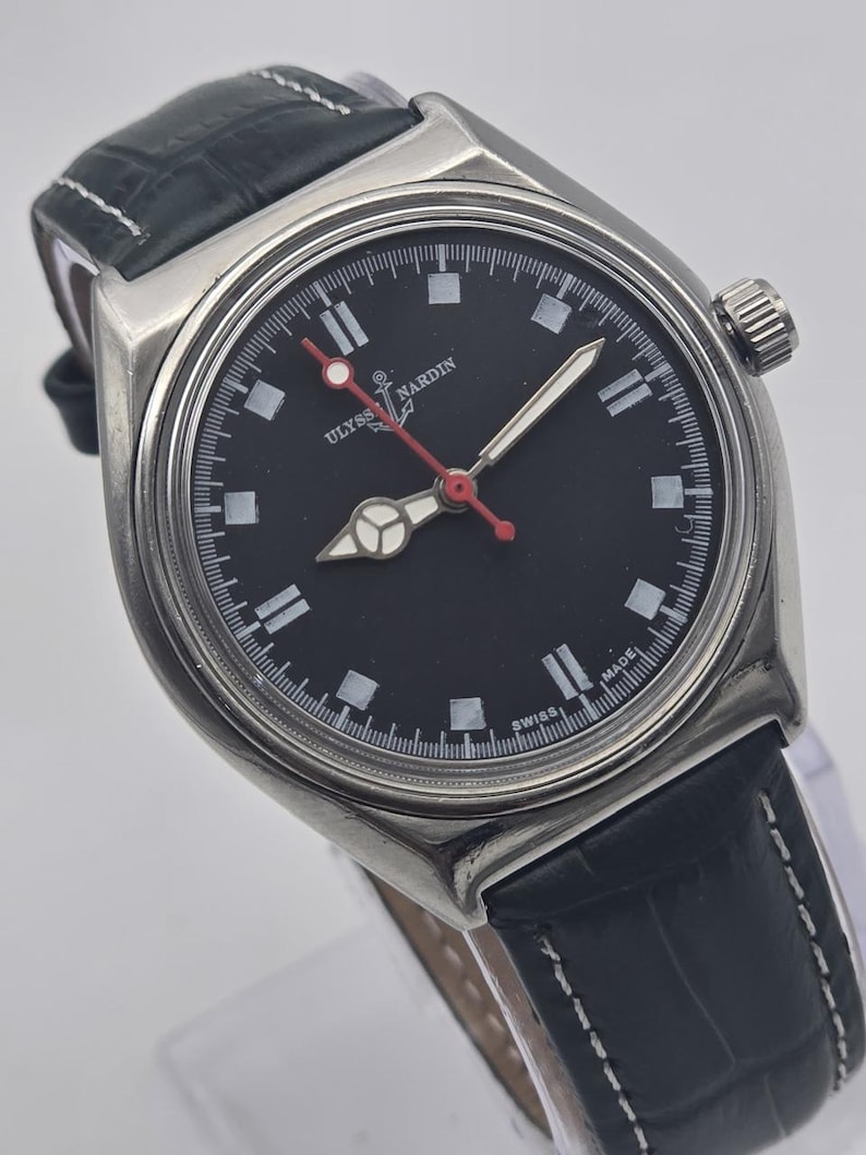 Puede incluir: Un reloj de pulsera de acero inoxidable con esfera negra y correa de cuero negro. El reloj presenta marcadores de hora blancos, segundero rojo y el texto "ULYSSES NARDIN SWISS MADE". Tiene un bisel plateado y una correa de cuero negro texturizada.