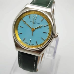 May include: A silver-toned wristwatch with a light blue face and gold-colored hands and accents. The watch has a green leather band with white stitching. The watch face has the text "ULYSSES NARDIN SWISS MADE".