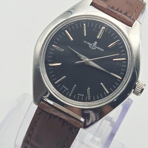 May include: A silver-toned Ulysse Nardin watch with a black face and brown leather band. The watch features gold-colored hour markers and hands. The band has a crocodile pattern. The watch is displayed on a clear stand.