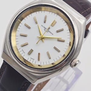 Pu&ograve; includere: Un orologio da polso Ulysse Nardin argentato con quadrante bianco, lancette e indici dorati e cinturino in pelle marrone. L'orologio ha una cassa rettangolare e un cinturino in pelle marrone scuro. Il quadrante riporta la scritta "Ulysse Nardin" e "Swiss Made".
