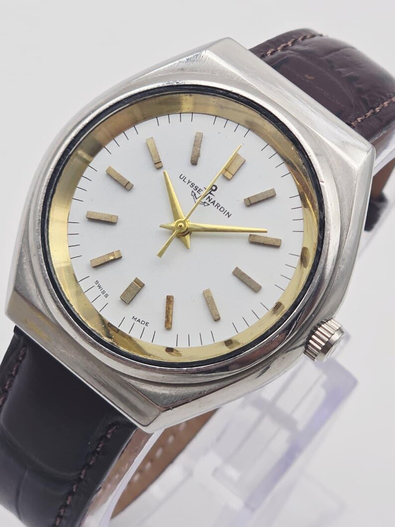 Pu&ograve; includere: Un orologio da polso Ulysse Nardin argentato con quadrante bianco, lancette e indici dorati e cinturino in pelle marrone. L'orologio ha una cassa rettangolare e un cinturino in pelle marrone scuro. Il quadrante riporta la scritta "Ulysse Nardin" e "Swiss Made".