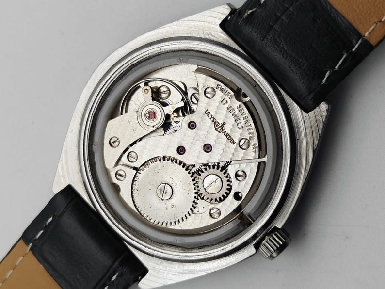 May include: Close-up of a vintage Ulysse Nardin watch with a silver-tone case and a black leather strap. The watch's intricate mechanical movement is visible through the clear back, showcasing gears, jewels, and the inscription "SWISS SEVENTEEN JEWELS".
