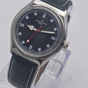 Puede incluir: Un reloj Ulysse Nardin de acero inoxidable con esfera negra, marcadores blancos y segundero rojo. El reloj tiene una correa de cuero negro con costuras blancas. El reloj se exhibe sobre un soporte transparente.