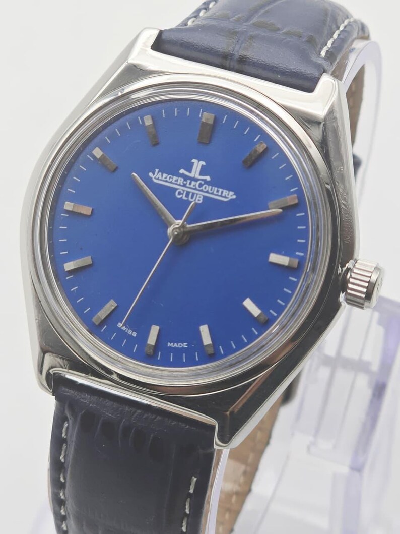May include: A stainless steel Jaeger-LeCoultre Club watch with a blue dial and a black leather strap. The watch features silver hands and markers, and the dial is marked with "JAEGER-LECOULTRE CLUB". The watch has a round face and a textured leather strap.