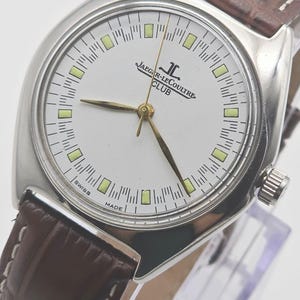 May include: A silver-toned Jaeger-LeCoultre Club watch with a white face and gold hands. The watch has a brown leather band. The face displays the brand name and the words "Swiss Made".