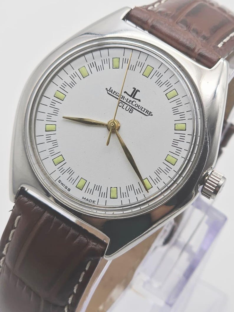 May include: A silver-toned Jaeger-LeCoultre Club watch with a white face and gold hands. The watch has a brown leather band. The face displays the brand name and the words "Swiss Made".
