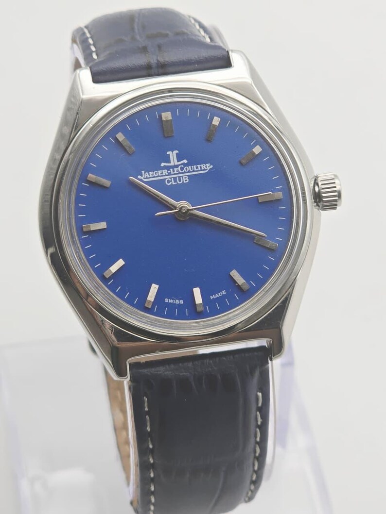 May include: A silver-toned Jaeger-LeCoultre Club watch with a blue dial and a black leather strap. The watch features silver hands and markers, and the words "SWISS MADE" are visible on the dial. The strap is approximately 8 inches long.