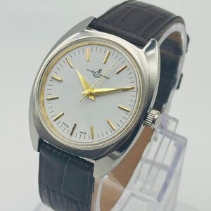 May include: A silver wristwatch with a light-coloured face and gold hands and markers. The watch features the text "ULYSSE NARDIN" and a dark brown leather strap. The watch is displayed on a clear stand.