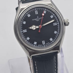 Puede incluir: Un reloj Ulysse Nardin de acero inoxidable con esfera negra, marcadores blancos y segundero rojo. El reloj tiene una correa de cuero con costuras blancas. El reloj se exhibe sobre un soporte transparente.