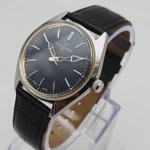 May include: A silver-toned Ulysse Nardin wristwatch with a dark blue face and black leather band. The watch has silver hands and hour markers. The band is textured with a crocodile pattern. The watch is displayed on a clear stand.
