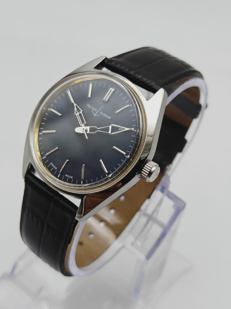 May include: A silver-toned Ulysse Nardin wristwatch with a dark blue face and black leather band. The watch has silver hands and hour markers. The band is textured with a crocodile pattern. The watch is displayed on a clear stand.