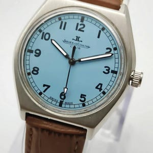 K&ouml;nnte beinhalten: Eine silberfarbene Armbanduhr mit hellblauem Zifferblatt und braunem Lederarmband. Das Zifferblatt zeigt schwarze Ziffern und den Markennamen "Jaeger-LeCoultre". Die Uhr hat ein rundes Zifferblatt und ein braunes Lederarmband.